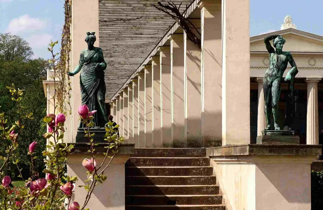 Dwie antyczne rzeźby w ogrodzie, w stylu klasycyzm, na tle kolumnady i kwitnącej magnolii.