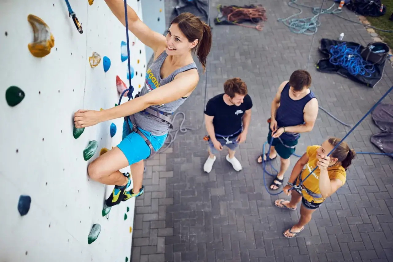 Frau beim **toprope klettern** an einer wei&szlig;en Kletterwand, gesichert von Freunden am Boden.