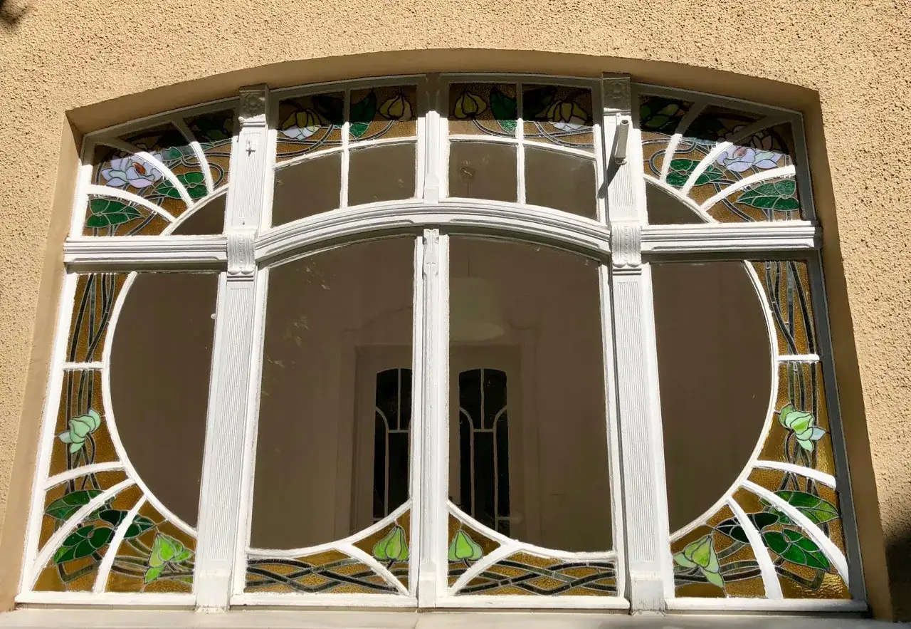 Magnifique **fen&ecirc;tre Art Nouveau** orn&eacute;e de vitraux aux motifs floraux et de lignes courbes, encadr&eacute;e de blanc sur un mur textur&eacute;.