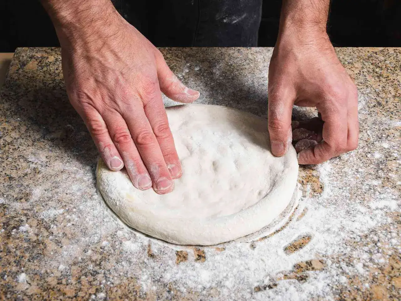 Dłonie formują okrągłe ciasto na pizzę, posypane mąką na granitowym blacie.