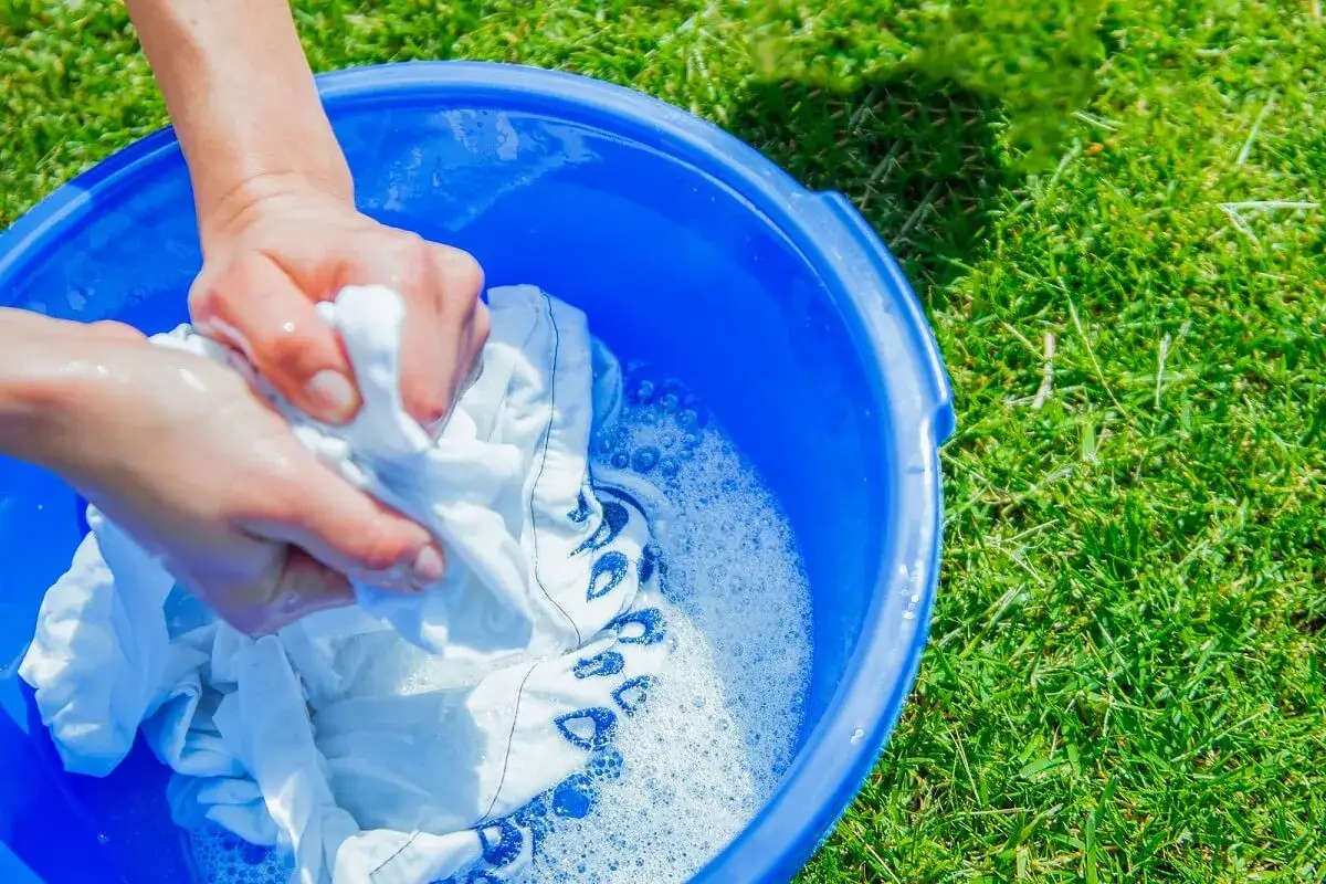 Manos lavan ropa blanca deste&ntilde;ida de azul en un cubo azul sobre c&eacute;sped verde.