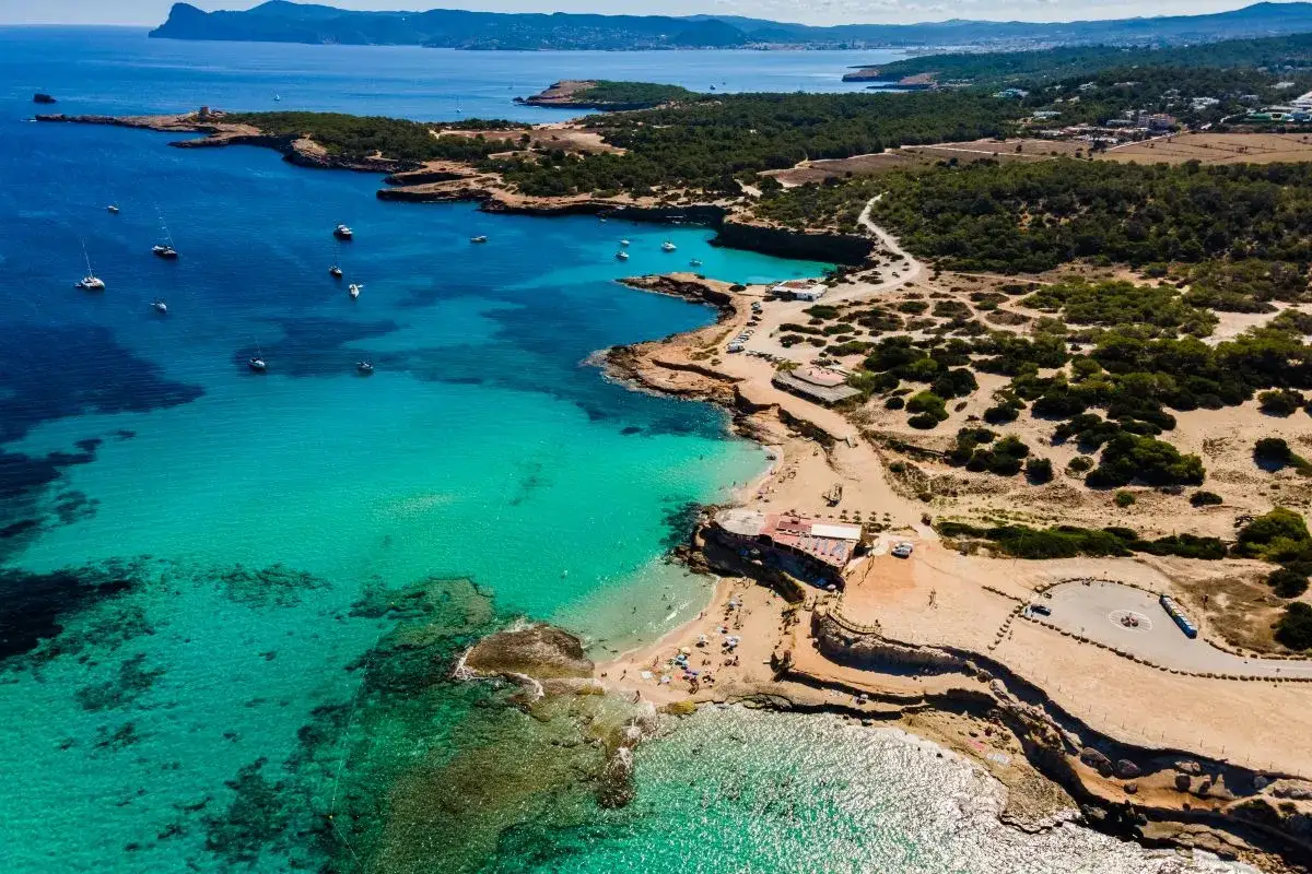 Blick auf eine Bucht mit türkisfarbenem Wasser und Segelbooten. Ein Strand mit Menschen und einem Gebäude, das an einen Ashram erinnert, liegt am Ufer. Ein malerischer Sonnenuntergang über Ibiza.