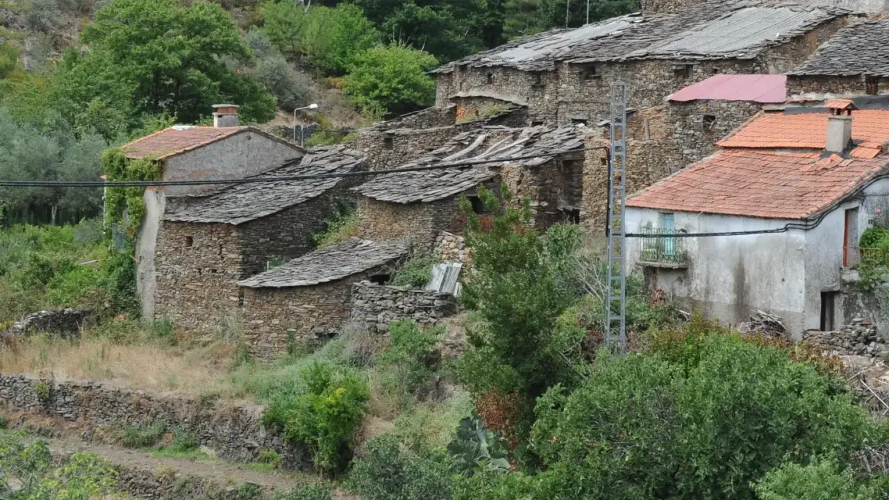 Casas de piedra con tejados de pizarra y teja roja en un pueblo de las Hurdes, rodeadas de vegetaci&oacute;n exuberante.