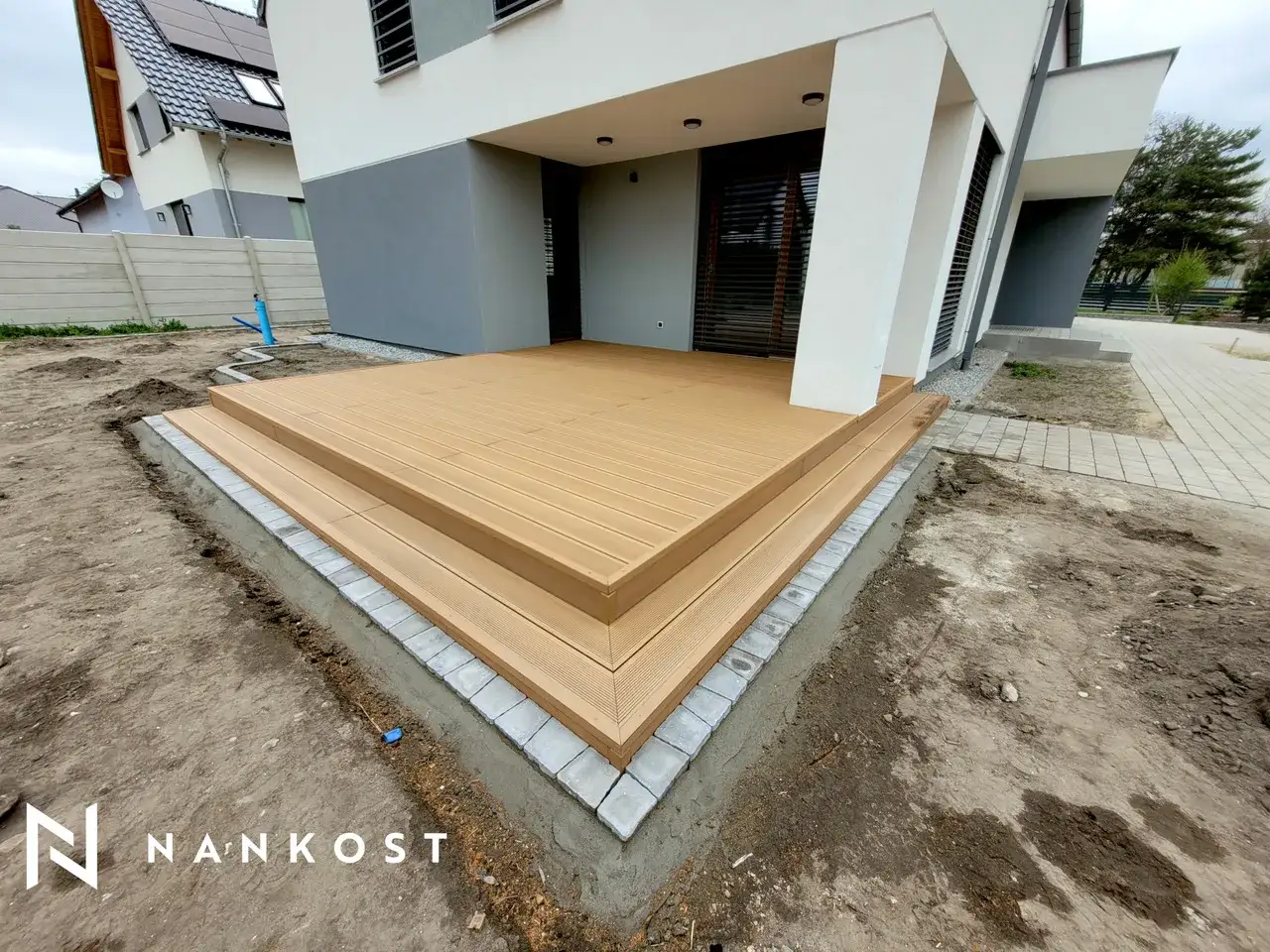Nowa deska kompozytowa na kostce brukowej, tworząca eleganckie schody i taras przy nowoczesnym domu.