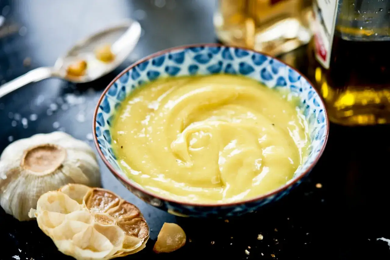 En skål med krämig aioli, en hel vitlök och en rostad vitlöksklyfta. Perfekt för ditt aioli recept.