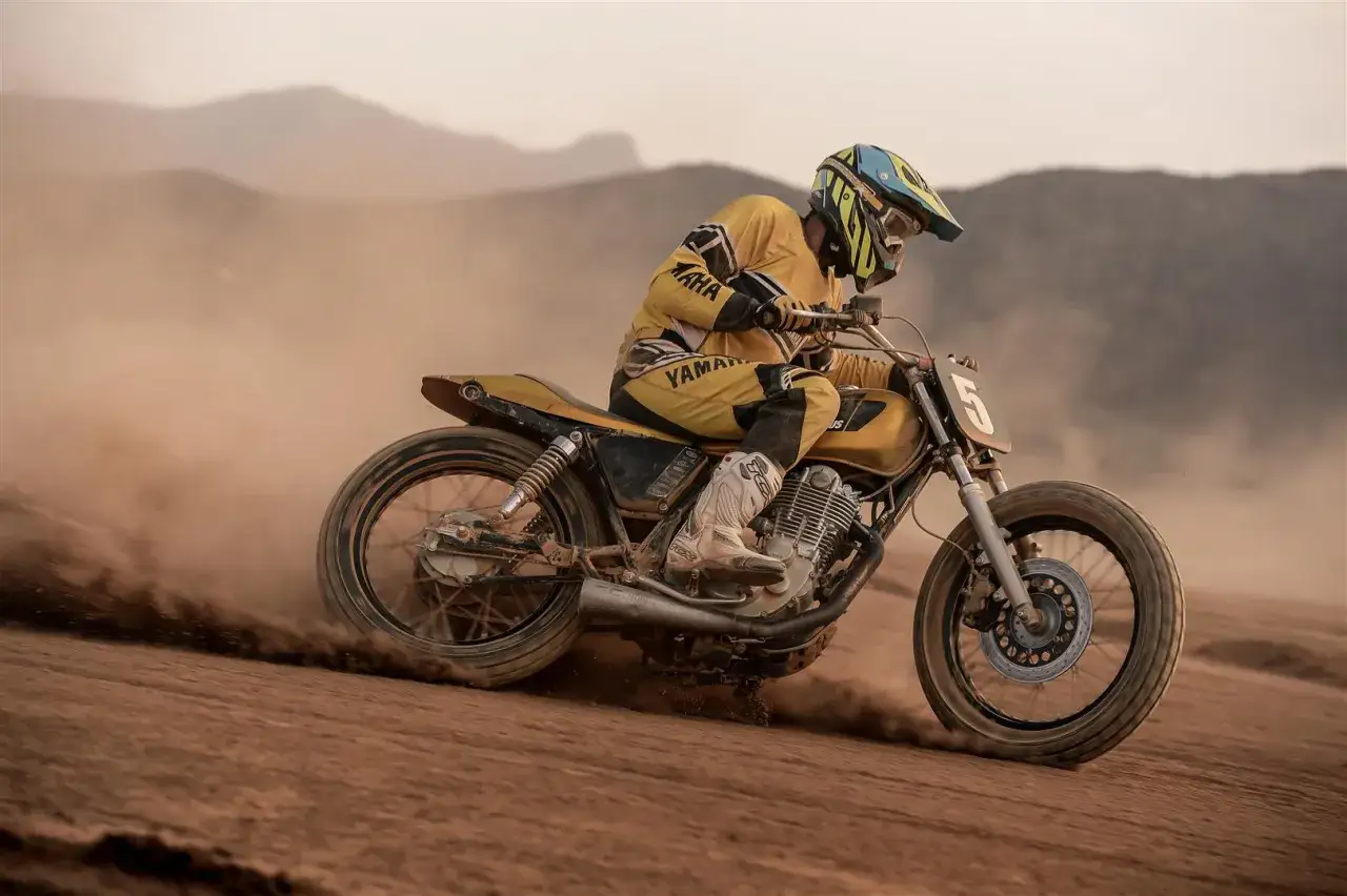 Piloto con casco y traje amarillo compite en una moto por una **dirt track**, levantando polvo en un paisaje desértico.
