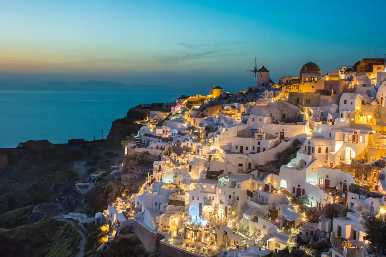 Oświetlone białe domy na zboczu klifu w Oia, Santorini, z widokiem na morze o zmierzchu. Wiatraki i kopuły kościołów dodają uroku. Idealne miejsce na wakacje, blisko lotnisko Santorini.