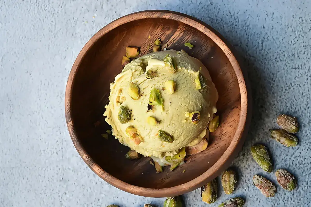 Cremoso helado de pistacho servido en un cuenco de madera, adornado con trozos de pistacho y acompañado de más pistachos enteros esparcidos.