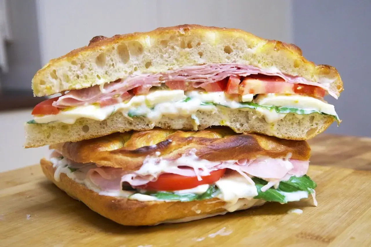 Dos bocadillos italianos apilados, rellenos de jamón, mozzarella, tomate y rúcula, sobre pan focaccia.