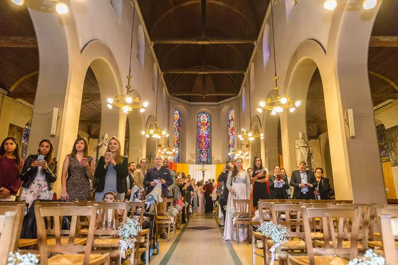 Des invités attendent dans une église, certains tenant leur téléphone, pour le cortege eglise mariage. Les vitraux illuminent la nef.