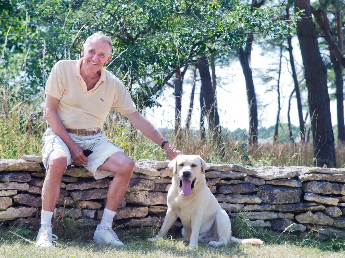 En leende man sitter på en stenmur med sin labrador. Kanske läser han ur sin **jan eliasson bok** i solen.
