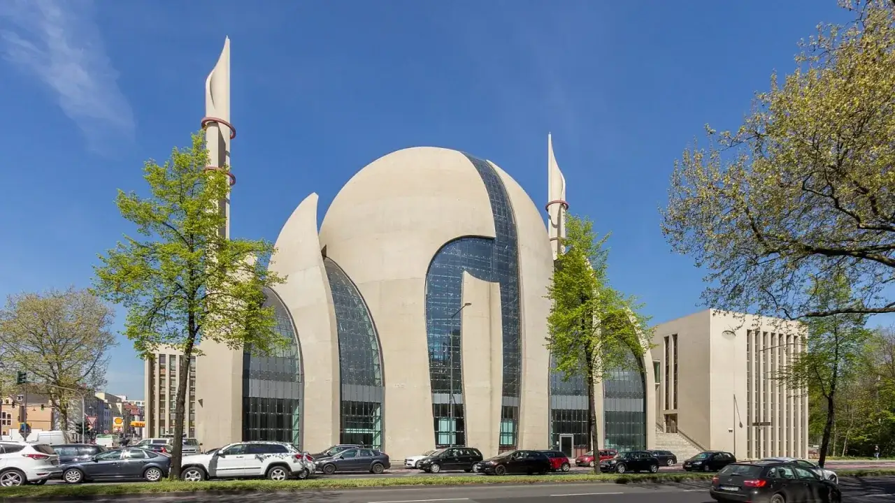 Nowoczesny meczet z minaretami, gdzie muezin wzywa do modlitwy. Na ulicy ruch samochodowy.