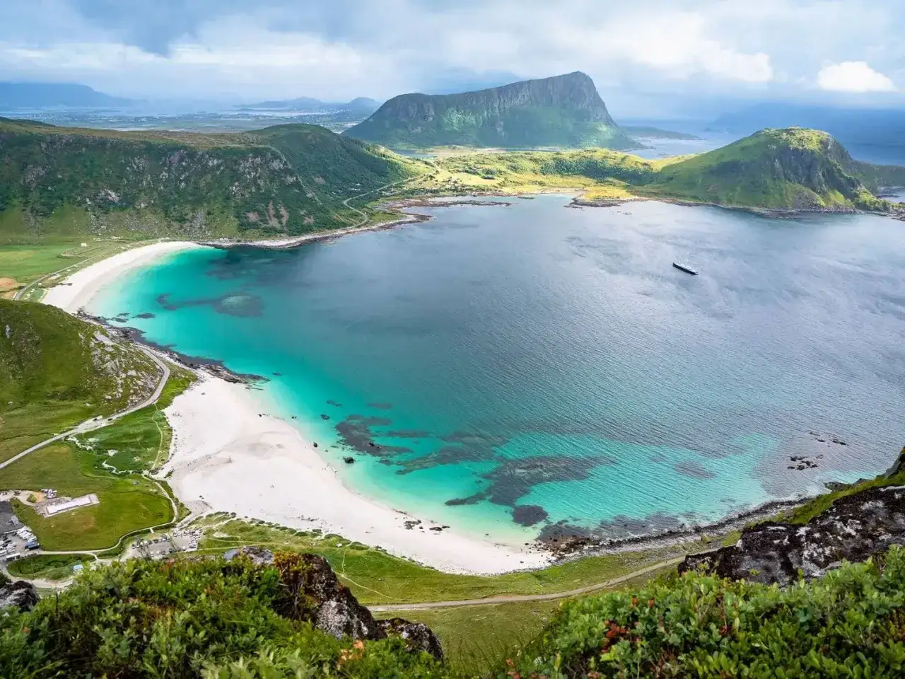 En dramatisk vy över en vit sandstrand och turkost vatten vid en Lofoten beach, omgiven av gröna berg under en molnig himmel.