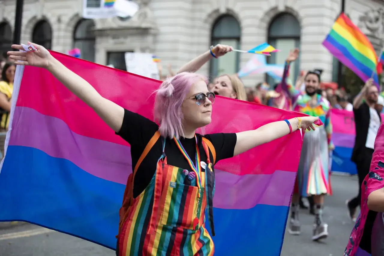Osoba z różowymi włosami dumnie niesie flagę biseksualnych. W tle widać innych uczestników parady.