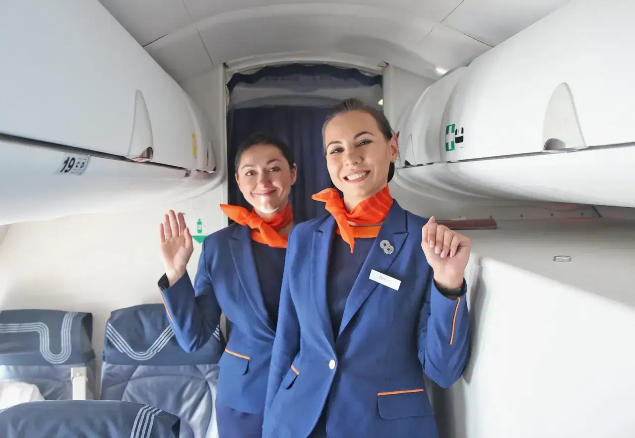 Dwie uśmiechnięte stewardessy w granatowych mundurach i pomarańczowych apaszkach. Spełniają wymagania, by zapewnić komfort podróży.