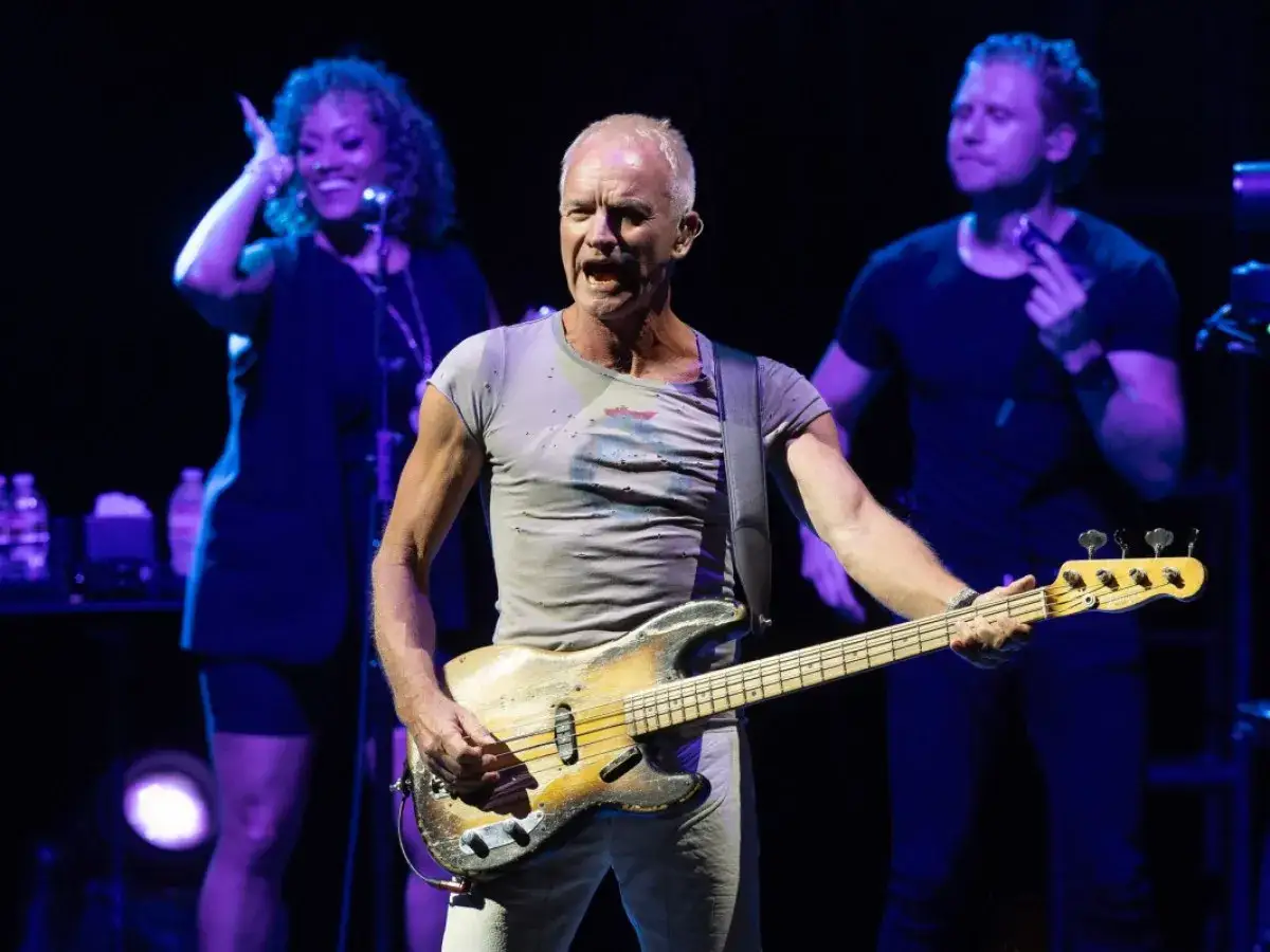 Sting, lider zespołu The Police, gra na gitarze basowej na koncercie. W tle wokalistka i gitarzysta.