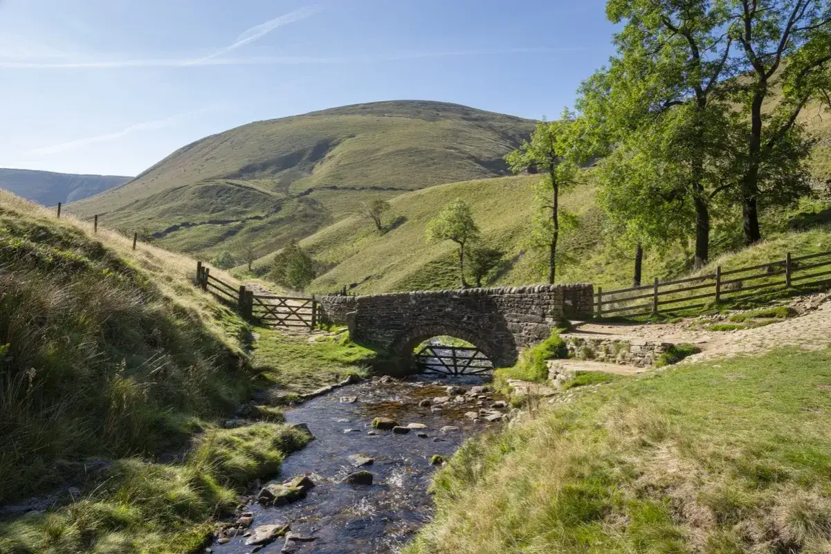 A stone bridge arches over a stream, inviting a peaceful hiking in England. Rolling green hills and wooden fences complete the idyllic scene.