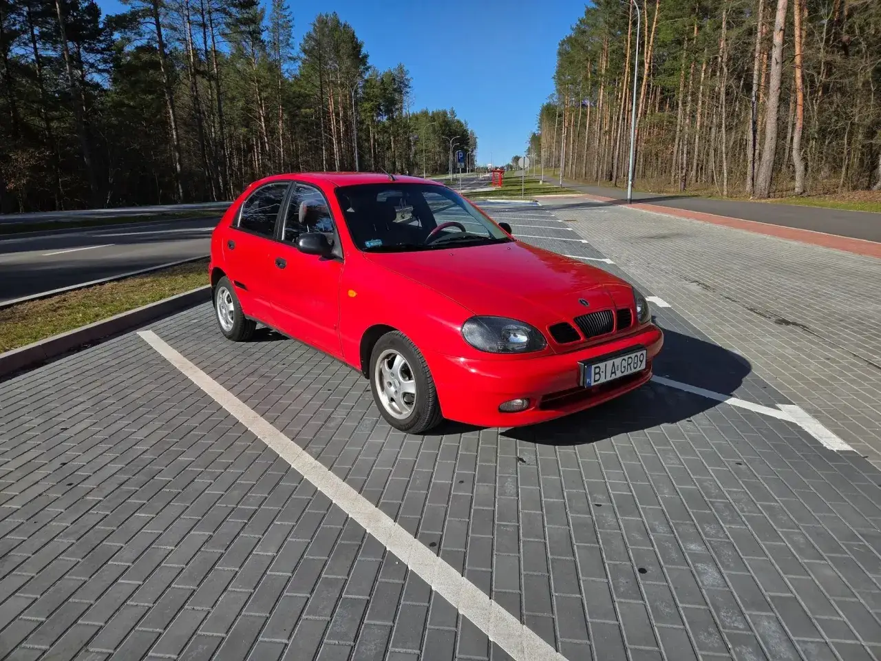 Czerwone Daewoo Lanos na parkingu z kostki brukowej, otoczonym lasem sosen.