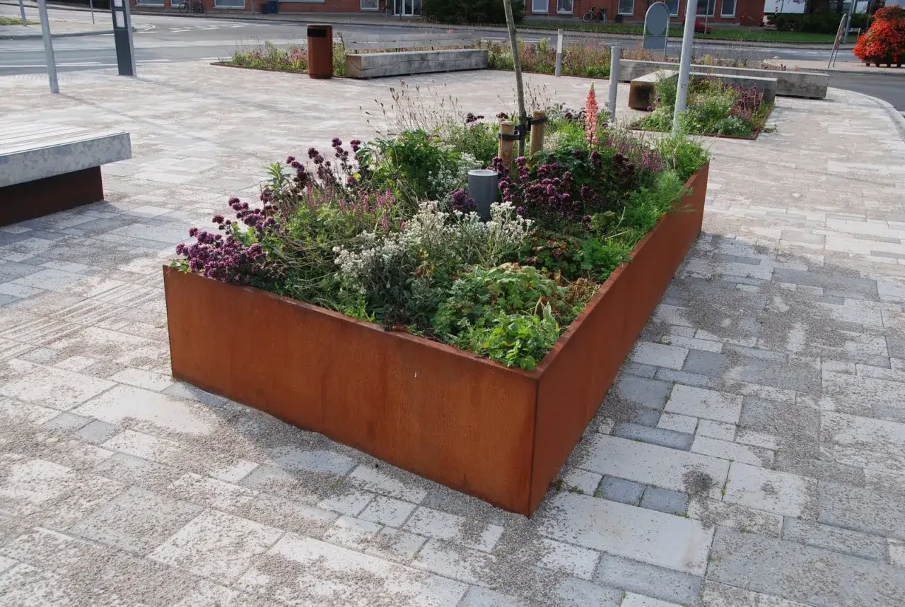 En rostig planteringslåda fylld med lila och vita blommor ger plantering inspiration i en stadsmiljö.