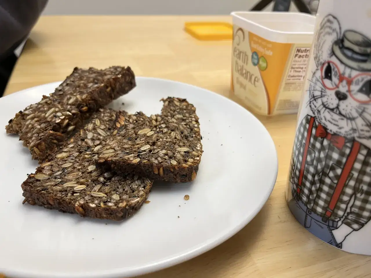 Drei Scheiben Vollkornbrot mit vielen Samen liegen auf einem Teller. Daneben eine Butterpackung und eine Tasse mit einem Kater. Ist Brot ungesund? Dieses hier wohl kaum!
