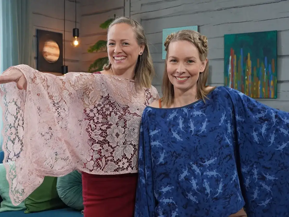 Två glada kvinnor visar upp sina nya sy blusar. Den ena bär en ljusrosa spetsblus, den andra en mörkblå med ödlemönster.
