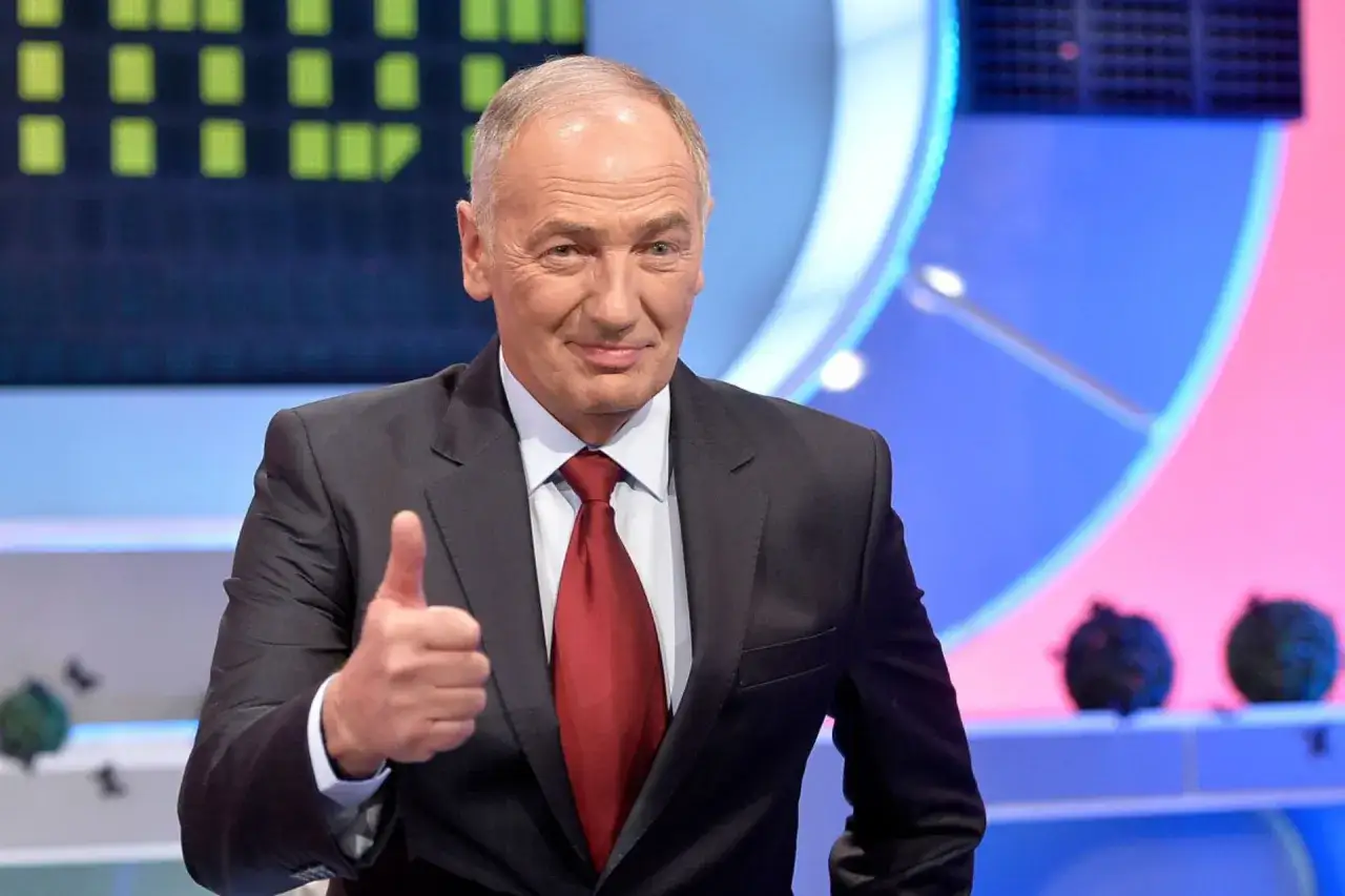 Uśmiechnięty mężczyzna w garniturze pokazuje kciuk w górę. W tle widać planszę z zielonymi polami, jak w teleturnieju Familiada.