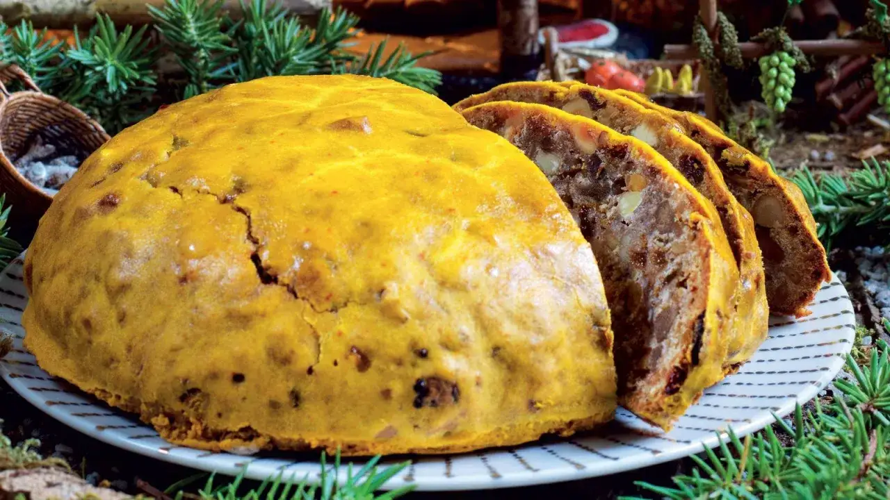 Pangiallo ricetta: un dolce natalizio dorato e ricco di frutta secca, tagliato a fette su un piatto a righe, circondato da rami di pino.