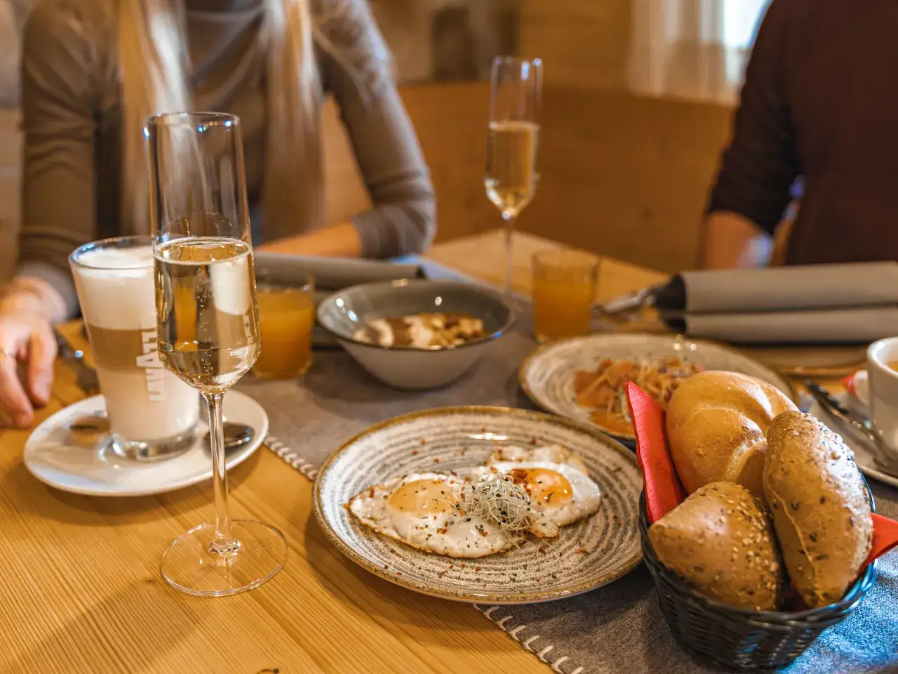 Fr&uuml;hst&uuml;ck mit Spiegeleiern, Brot und Sekt. Ein Glas Latte Macchiato und Orangensaft stehen bereit.