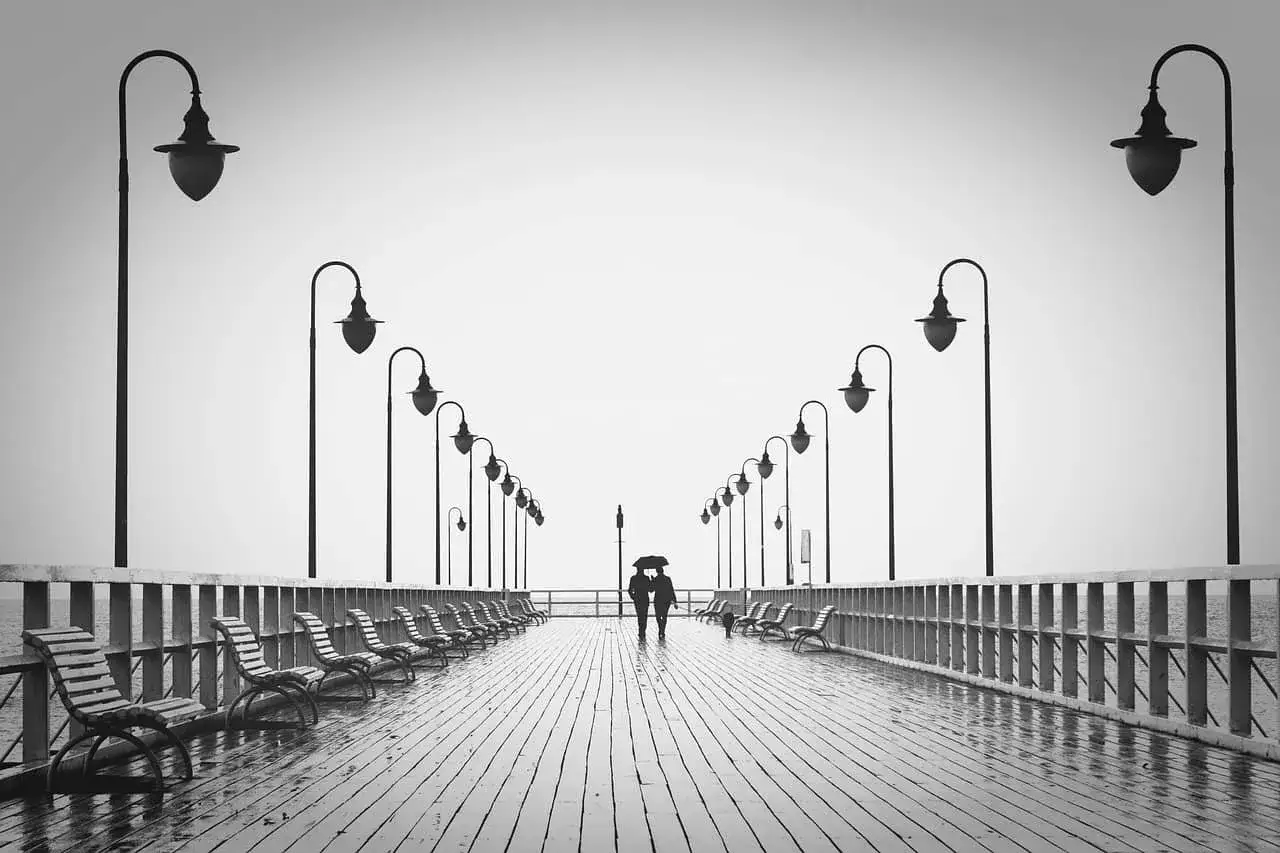 Pareja bajo paraguas en un muelle desolado, reflejando la decepci&oacute;n de pareja en un d&iacute;a gris.