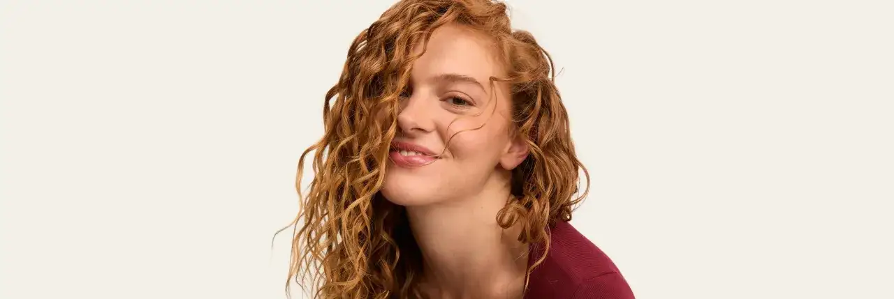 Une jeune femme aux cheveux roux bouclés, un sourire espiègle, une mèche de cheveux formant un joli noeud boucle sur sa joue.
