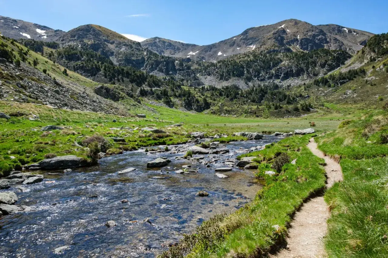 Sendero junto a un río pedregoso en el **Vall d'Incles**, rodeado de prados verdes y montañas boscosas bajo un cielo azul.