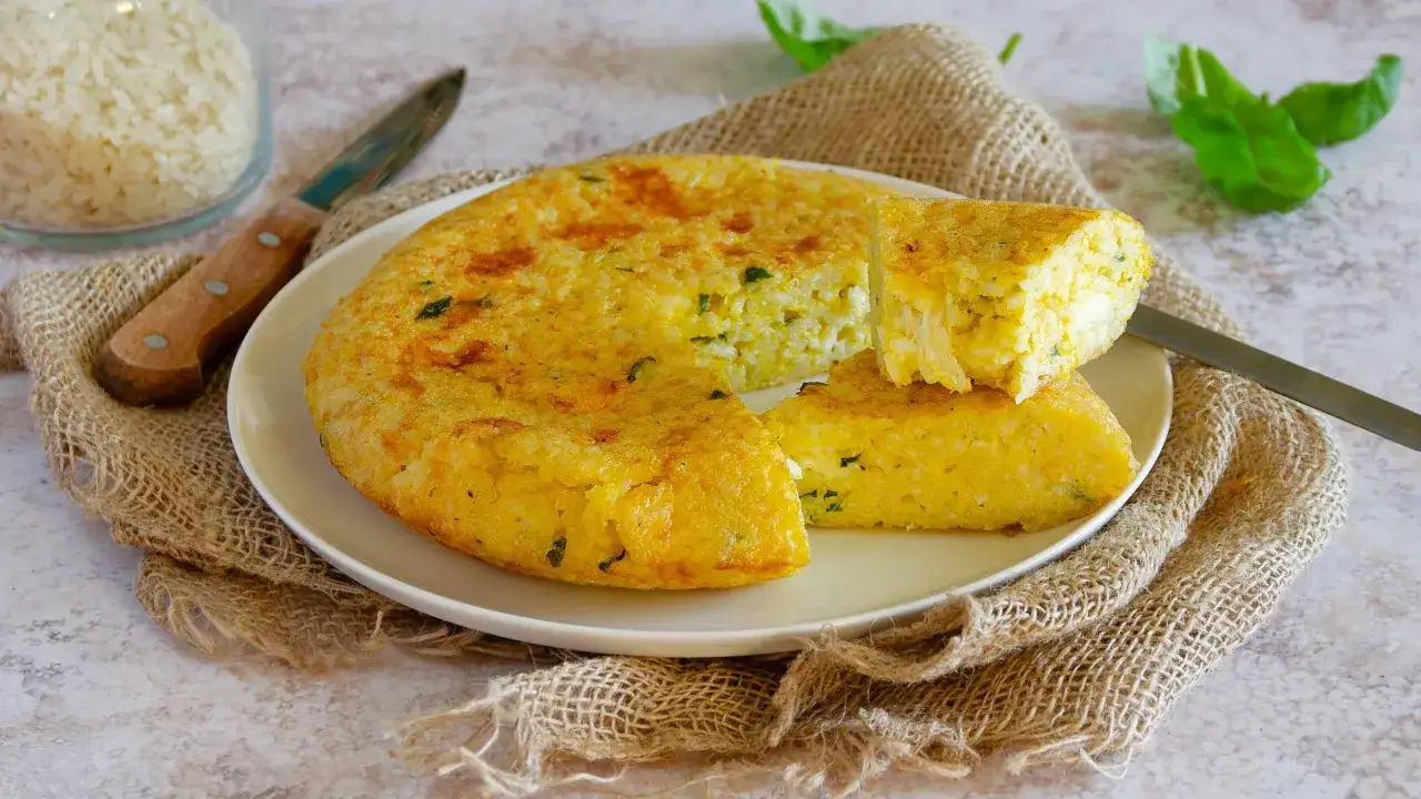 Una gustosa frittata di riso dorata, con erbe aromatiche, servita su un piatto bianco con un coltello e un barattolo di riso sullo sfondo.