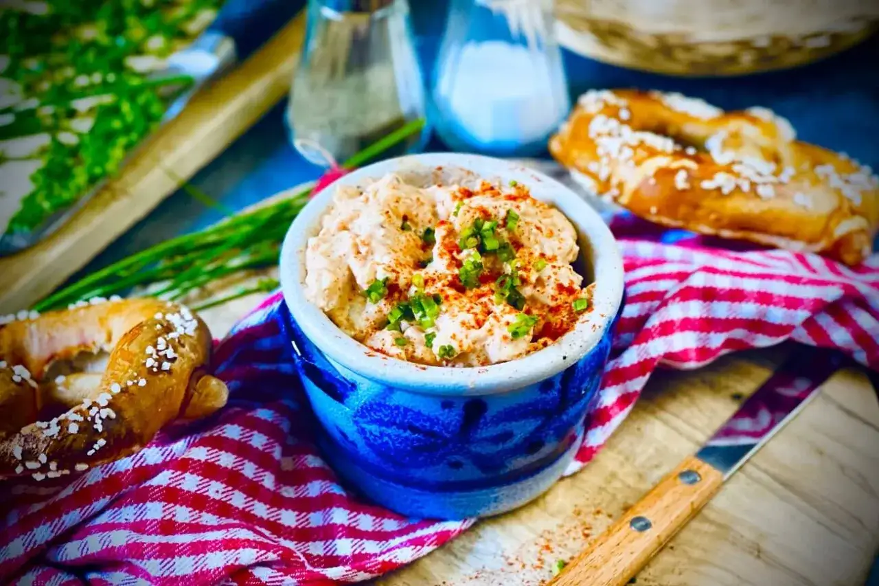 Herzhaftes Obazda-Rezept in einem blauen Steingut-Napf, garniert mit Schnittlauch und Paprikapulver, daneben Brezeln und ein Messer.