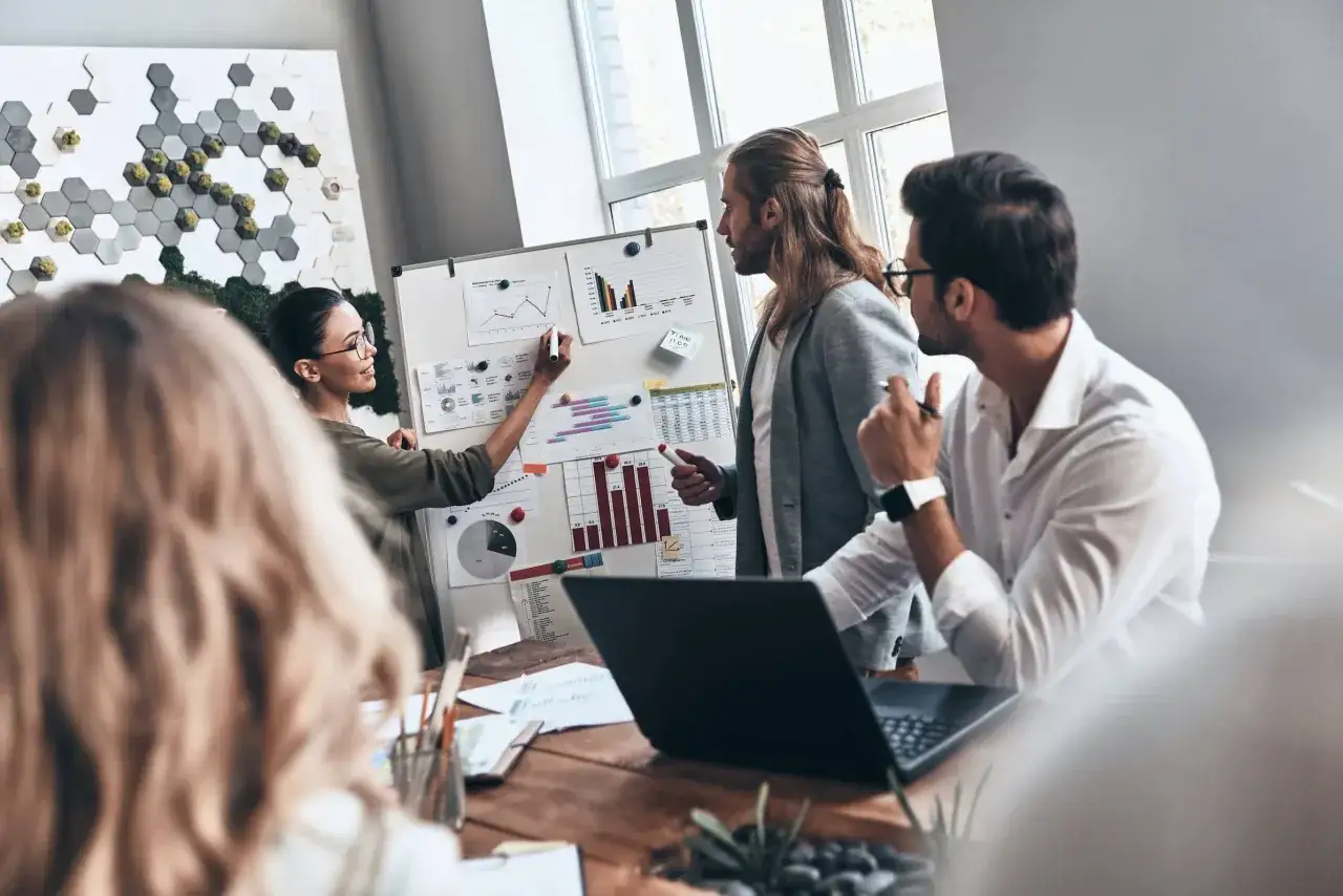 A team collaborates, discussing charts on a whiteboard. This scene shows how to be more authentic in business by openly sharing ideas and data.