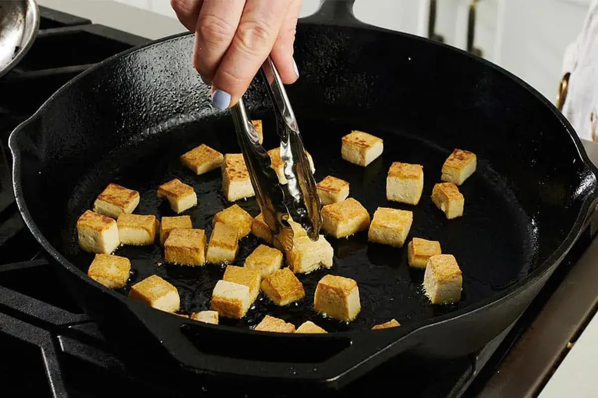 Cubos de tofu crujiente dorándose en una sartén de hierro fundido. Unas pinzas voltean un trozo para asegurar una cocción uniforme.