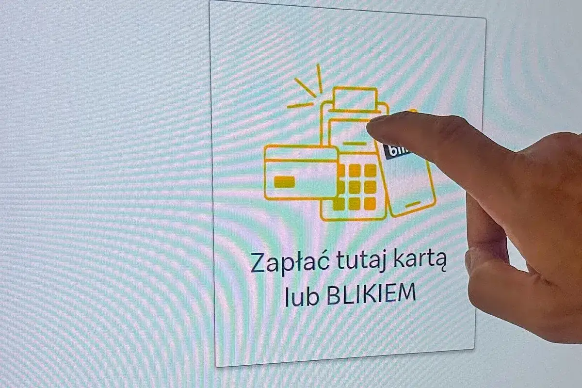 Palec wskazuje na ekran z grafiką terminala płatniczego, karty i telefonu. Napis 