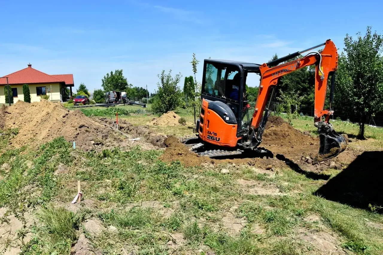 Mini koparka pracuje na budowie, gdzie prowadzony jest nadzór archeologiczny. W tle dom i drzewa.