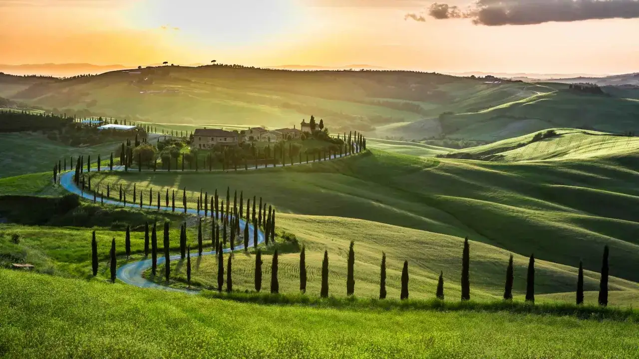 Dolce collina toscana al tramonto, con una strada tortuosa fiancheggiata da cipressi, perfetta per i tuoi itinerari in Toscana in macchina.