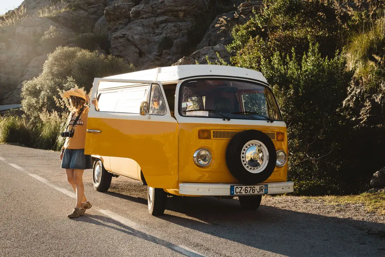 Une jeune femme avec un appareil photo et un chapeau de paille s'appuie sur la porte ouverte d'un van jaune, pr&ecirc;te pour une aventure de **Majorque en van**.