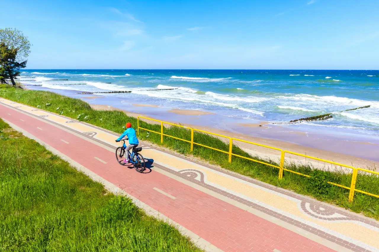 Kobieta w niebieskiej kurtce i czerwonej czapce jedzie rowerem po malowniczej ścieżce rowerowej wzdłuż plaży. To idealne miejsce na odkrywanie tras rowerowych Trójmiasta.