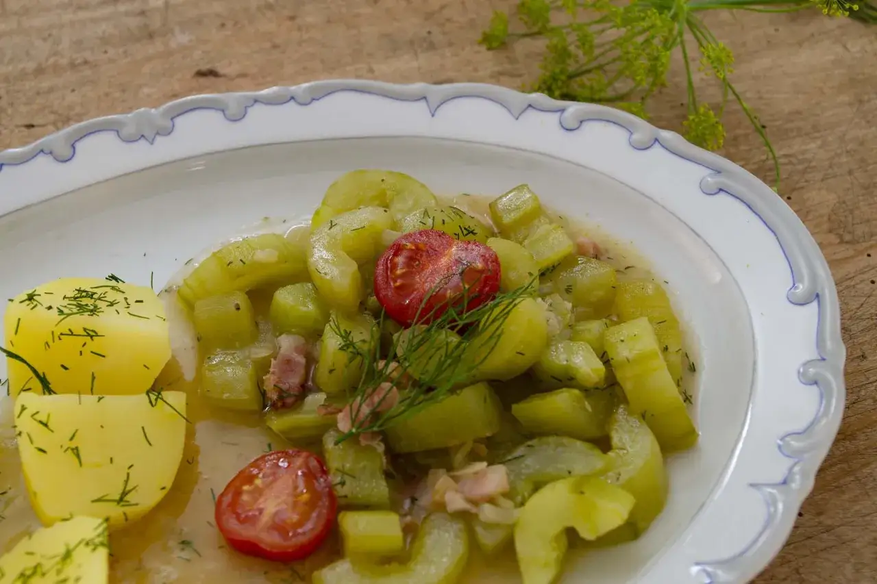 Schmorgurken sind hellgrün, in Scheiben und Würfel geschnitten, mit Tomaten und Speck auf einem Teller mit Kartoffeln.