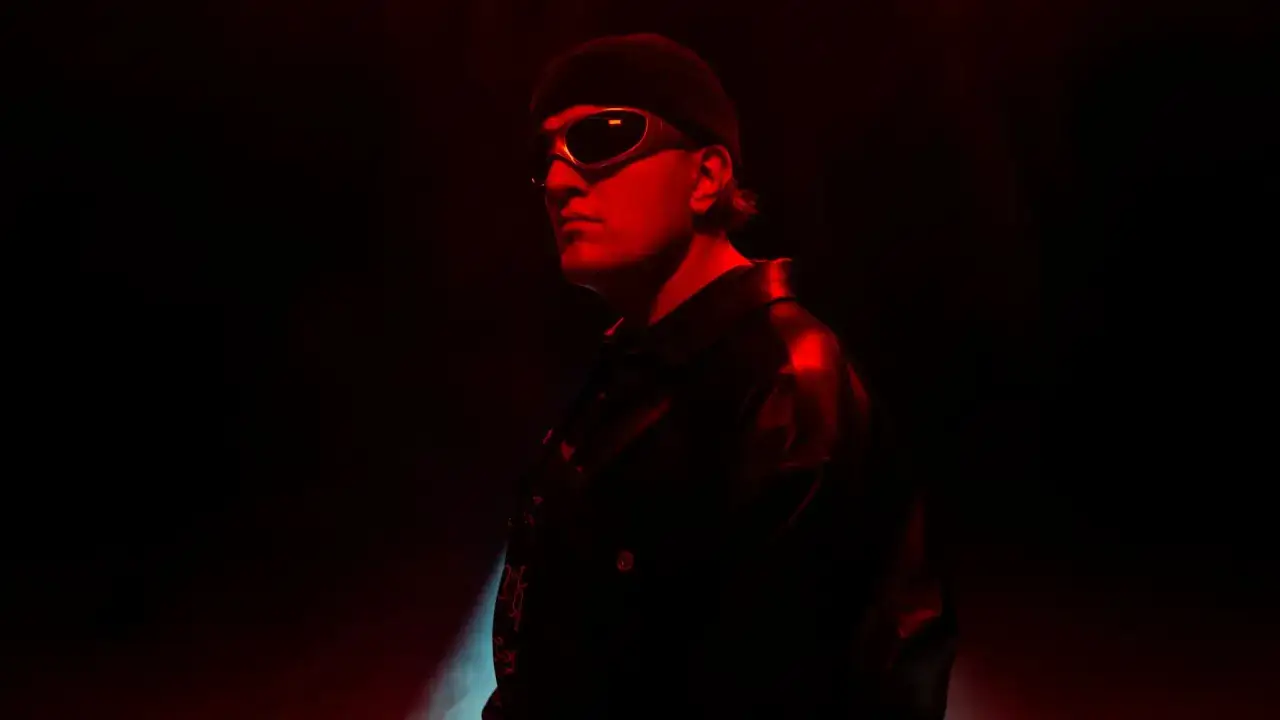Gigi D'Agostino w okularach i czapce, w czerwonym świetle. Mimo choroby, jego styl wciąż przyciąga uwagę.