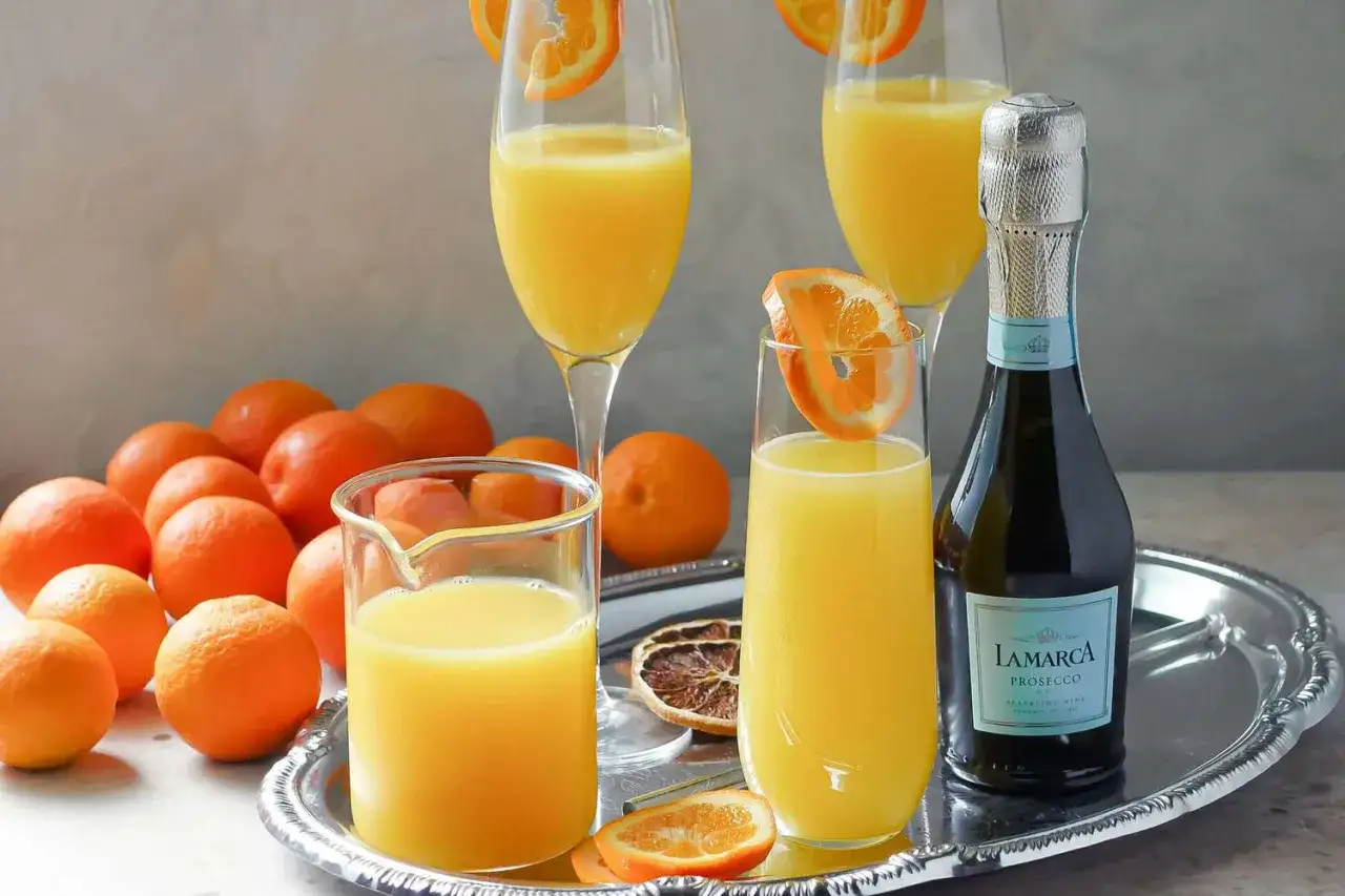 Orzeźwiający drink z prosecco i sokiem pomarańczowym, ozdobiony plasterkiem pomarańczy. Obok butelka prosecco i świeże pomarańcze.