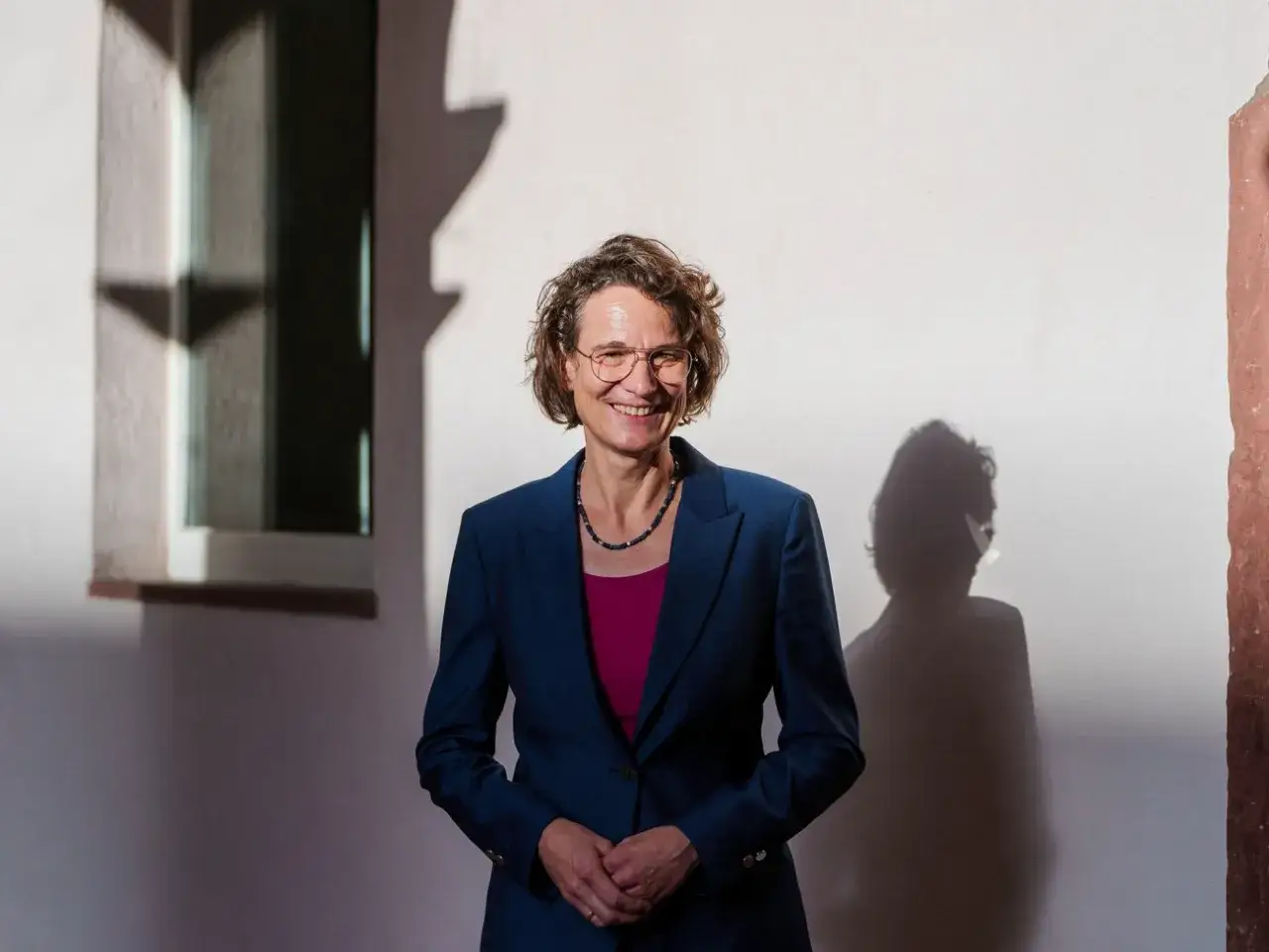 Christiane Tietz lächelt in die Kamera, gekleidet in einen blauen Anzug. Ihr Schatten wirft sich an die Wand.