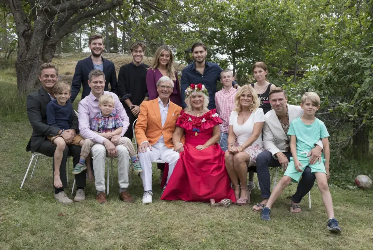 Stor familj samlad utomhus. I mitten sitter en kvinna med blomsterkrans, omgiven av sina barn och barnbarn. En av dem &auml;r Hans Wahlgrens barnbarn.