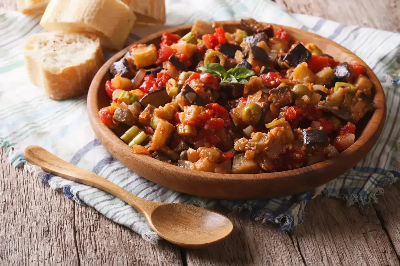 Un plat de caponata sicilienne, un mélange coloré d'aubergines, tomates, olives et céleri, servi dans un bol en bois avec du pain frais et une cuillère.