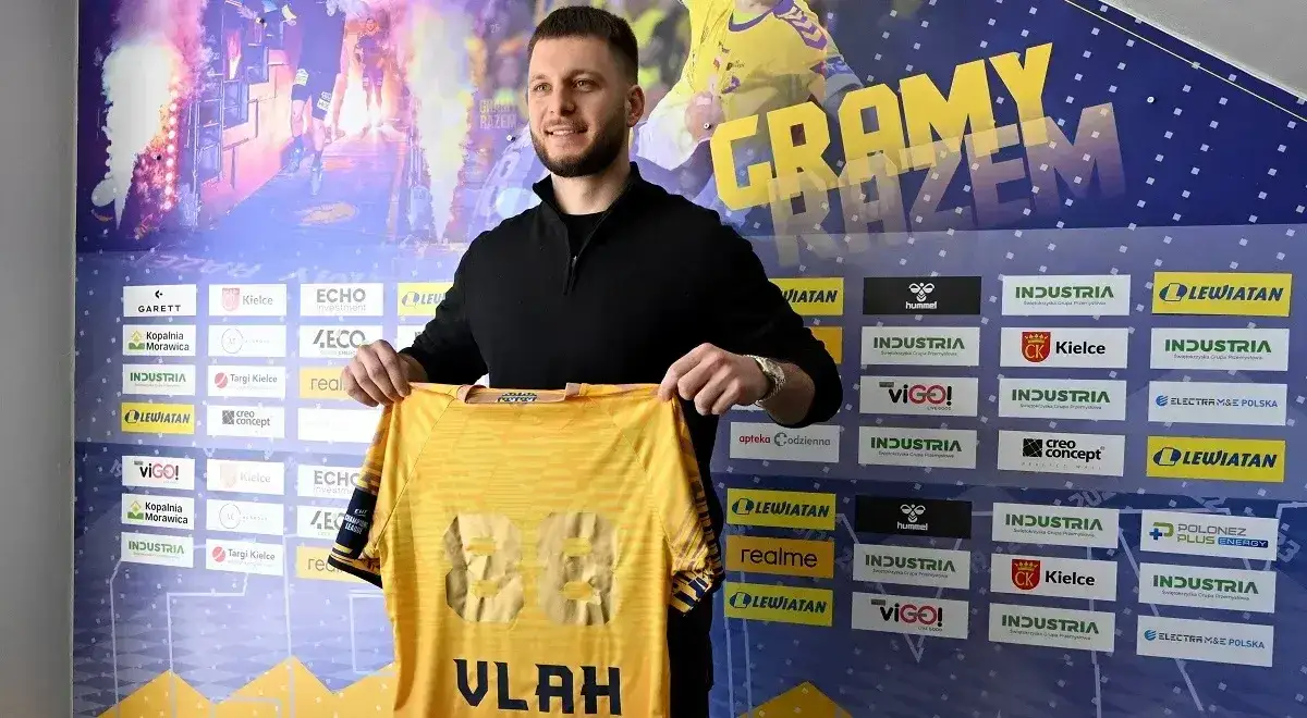 Aleks Vlah prezentuje nową koszulkę z numerem 88. Uśmiechnięty sportowiec pozuje na tle baneru z logo klubu i sponsorów.
