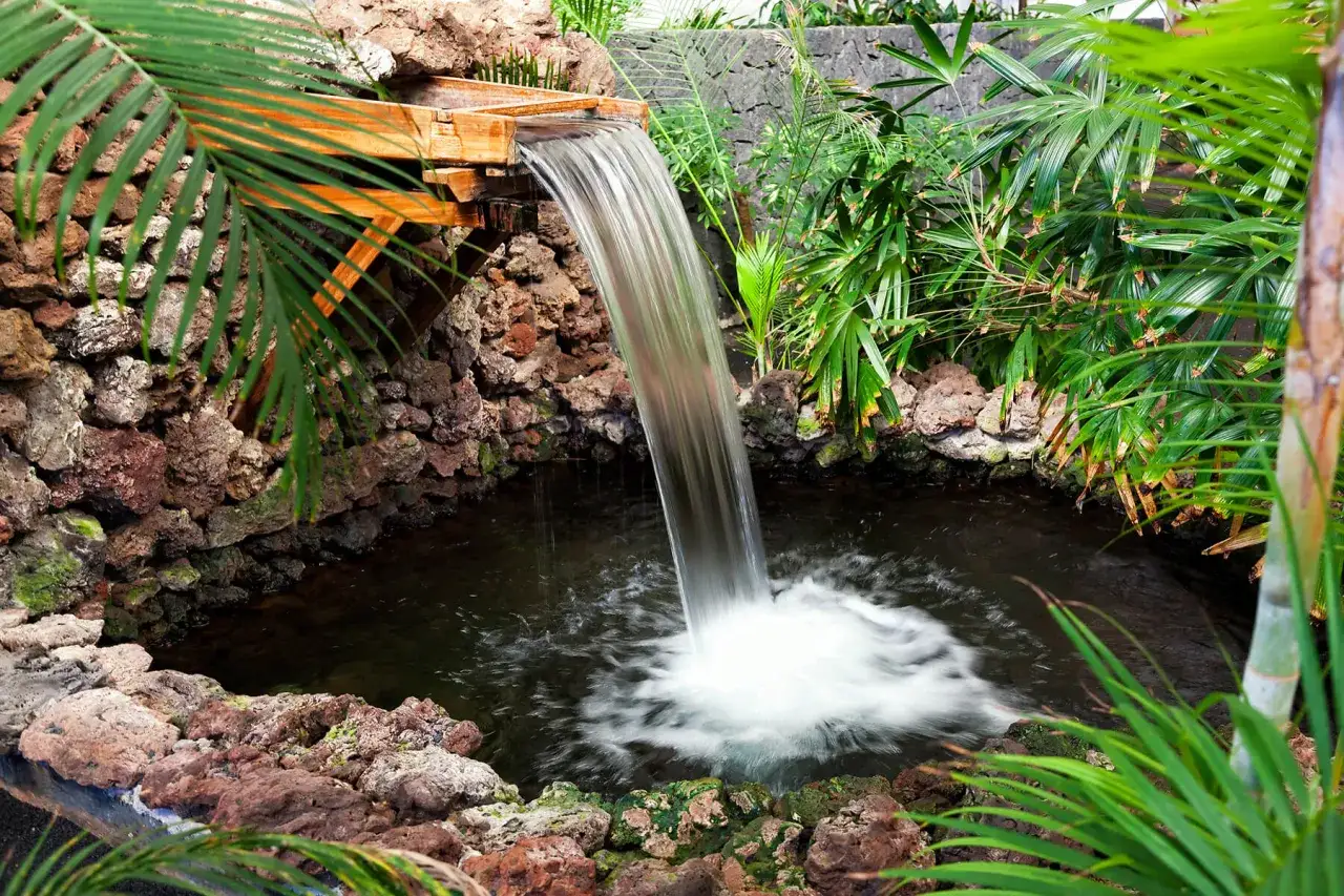 Ein kleiner Wasserfall aus Holz pl&auml;tschert in einen Teich, umgeben von Felsen und &uuml;ppiger Vegetation. Tolle Wasserfall Garten Ideen f&uuml;r eine Oase der Ruhe.