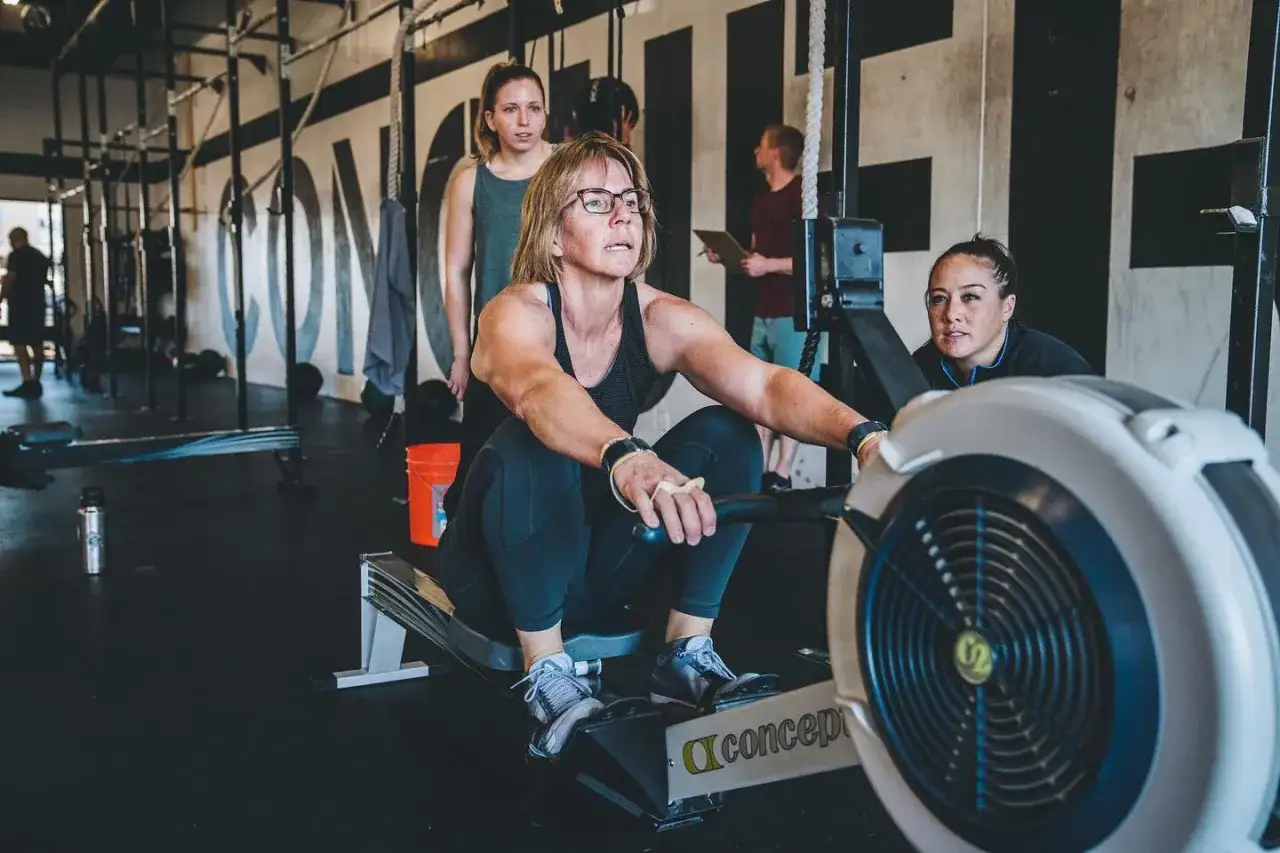 Kobieta w okularach wykonuje trening crossfit na wioślarzu, wspierana przez dwie inne kobiety.