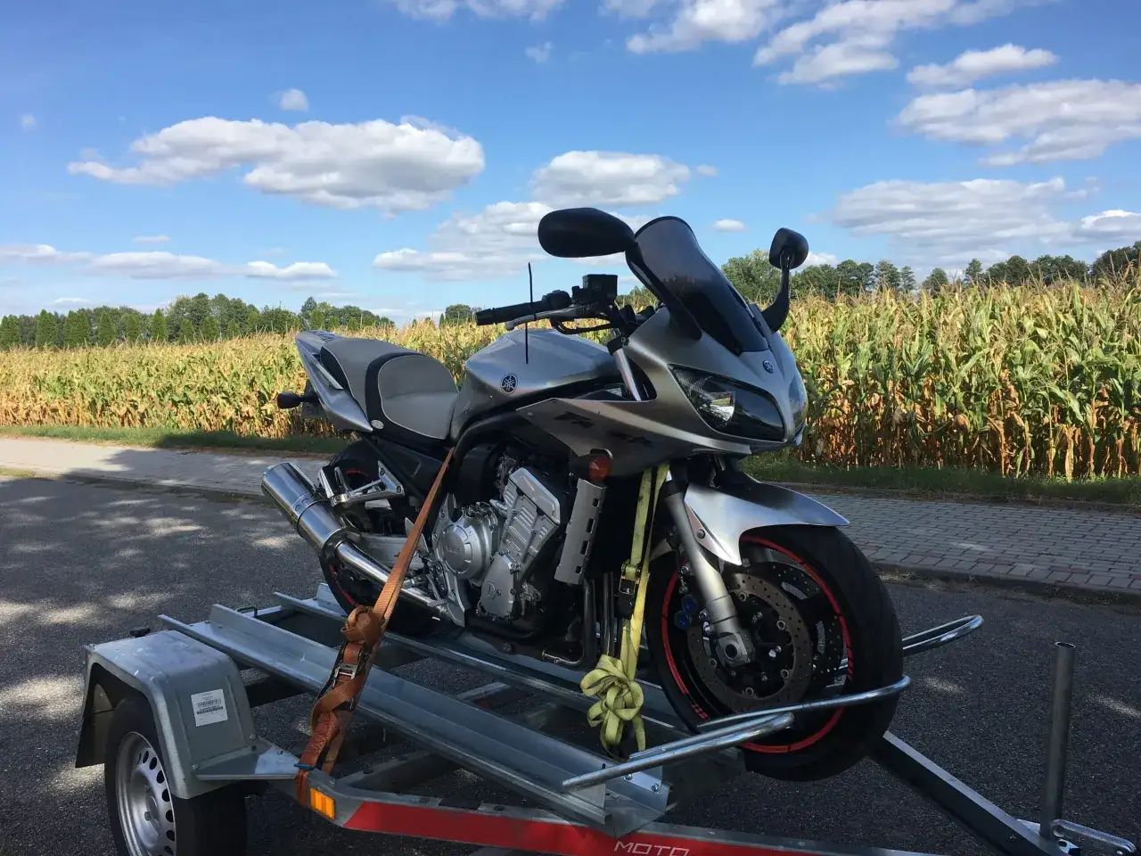 Srebrny motocykl Yamaha na przyczepce, gotowy do transportu. W tle pole kukurydzy i błękitne niebo. Myślę o akcyzie na motocykle.